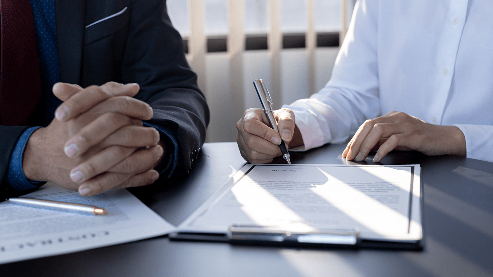 dame underskriver et fysisk dokument med en mand sidder og fletter hans fingre. Billedet er lyst og indbydende. billedet er zoomet ind så det kun er hænderne, bordet og papiret man kan se