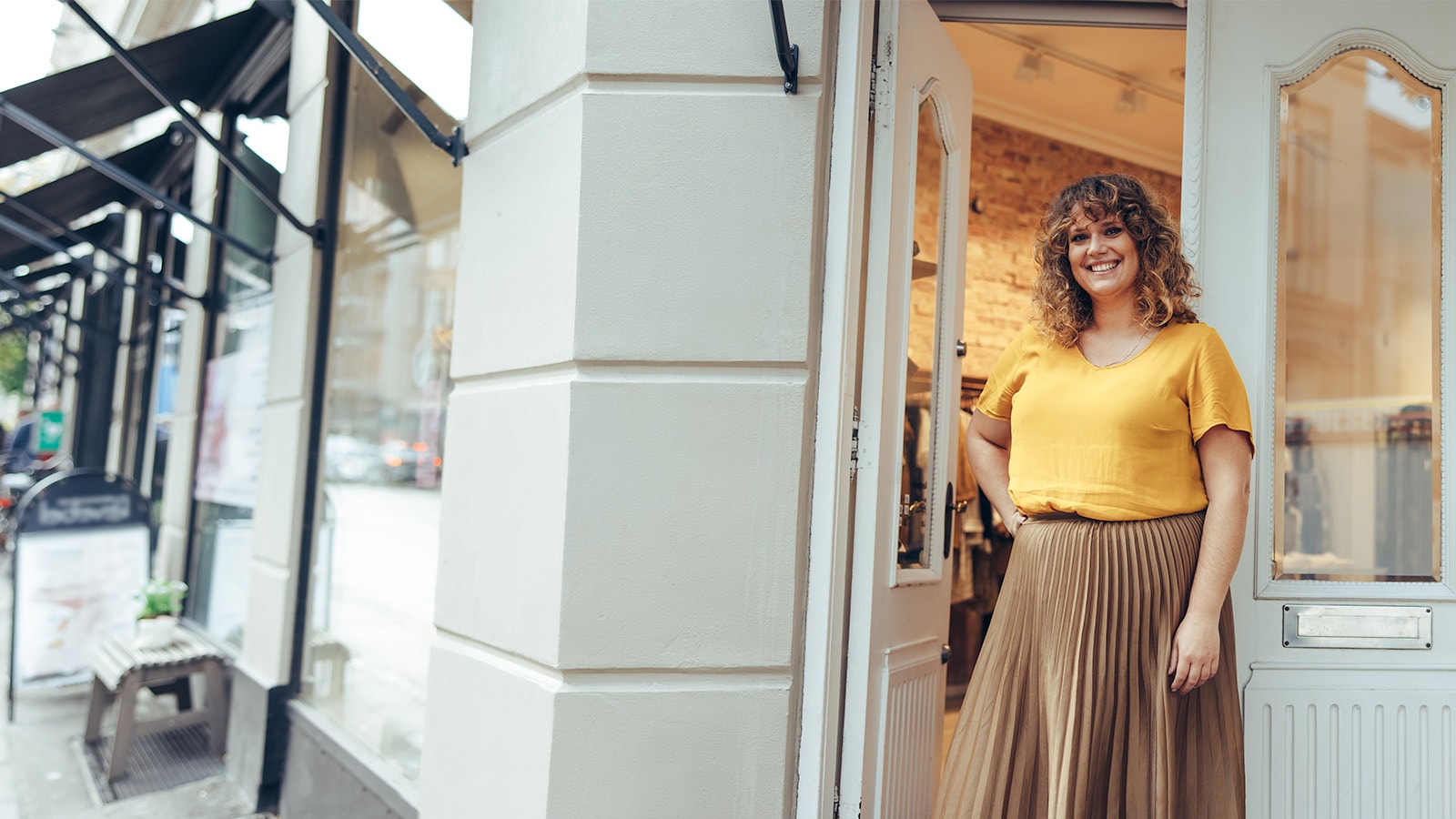 Dame i gul tshirt står ved indgangen til hendes tøjbutik og smiler ud til kameraet