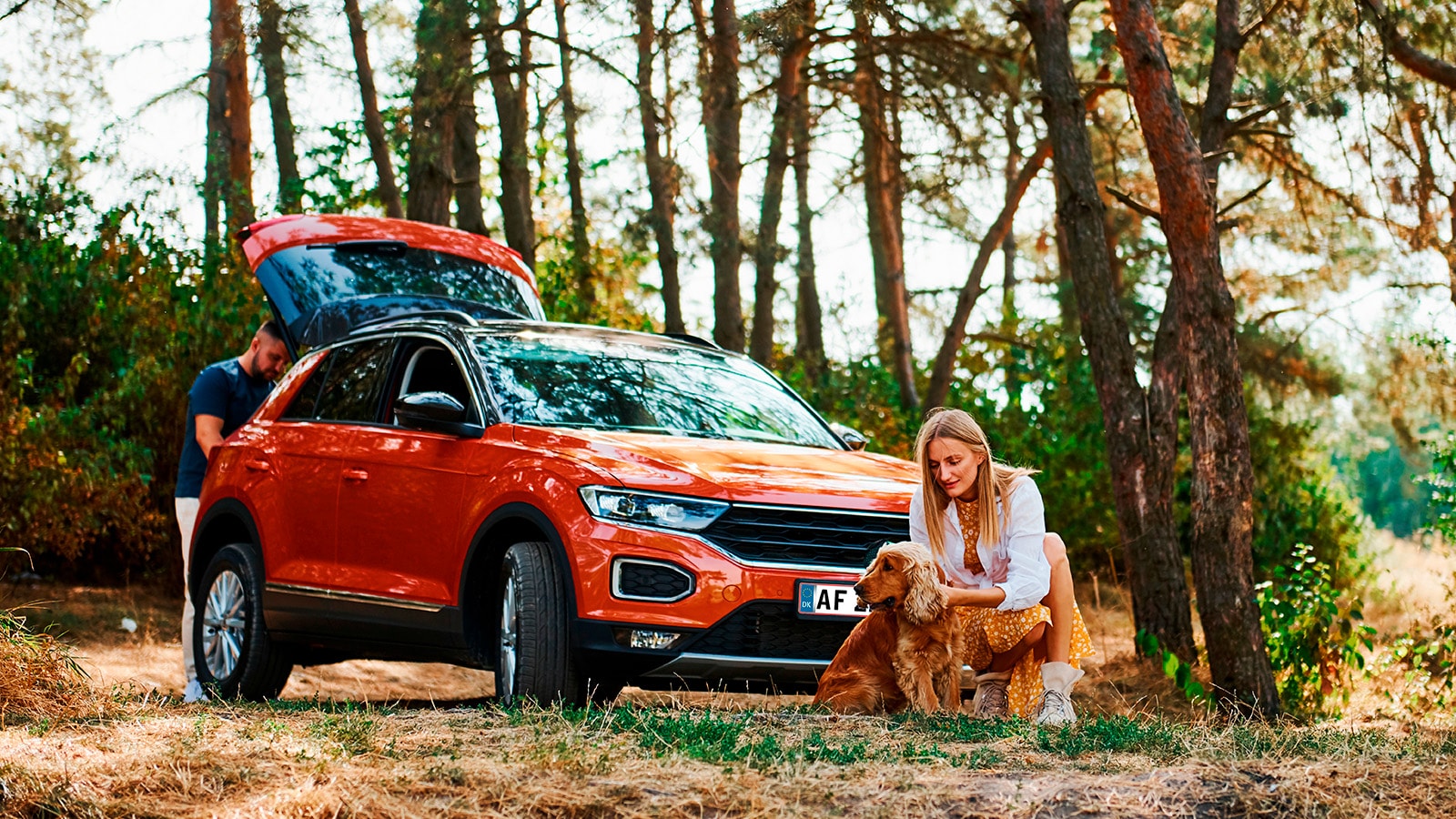 Rød bil ude i en skov med bagagerummet åbent, en dame og hendes hund tager en pause