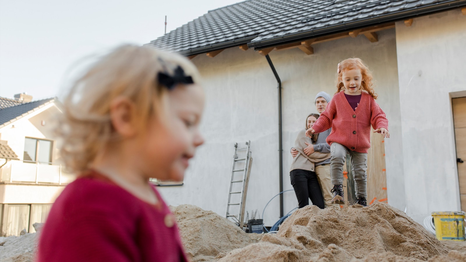 2 børn leger på en jordbunke hvor de bygger hyt hus. Far og mor står i baggrunden og kigger på