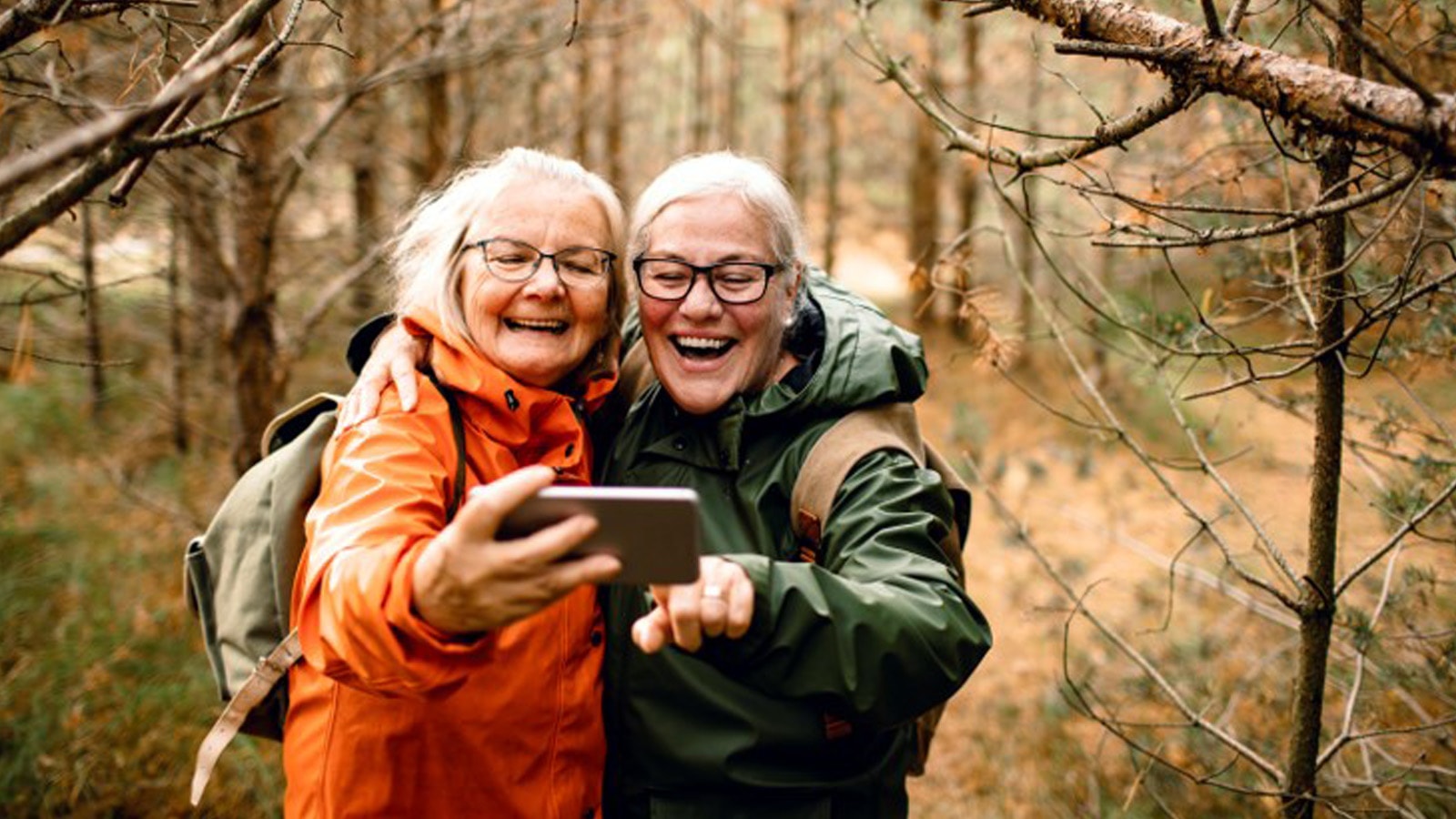 To ældre kvinder på tur i skoven en efterårsdag med rygsæk og jakke på. tager en selfie med den enes mobiltelefon