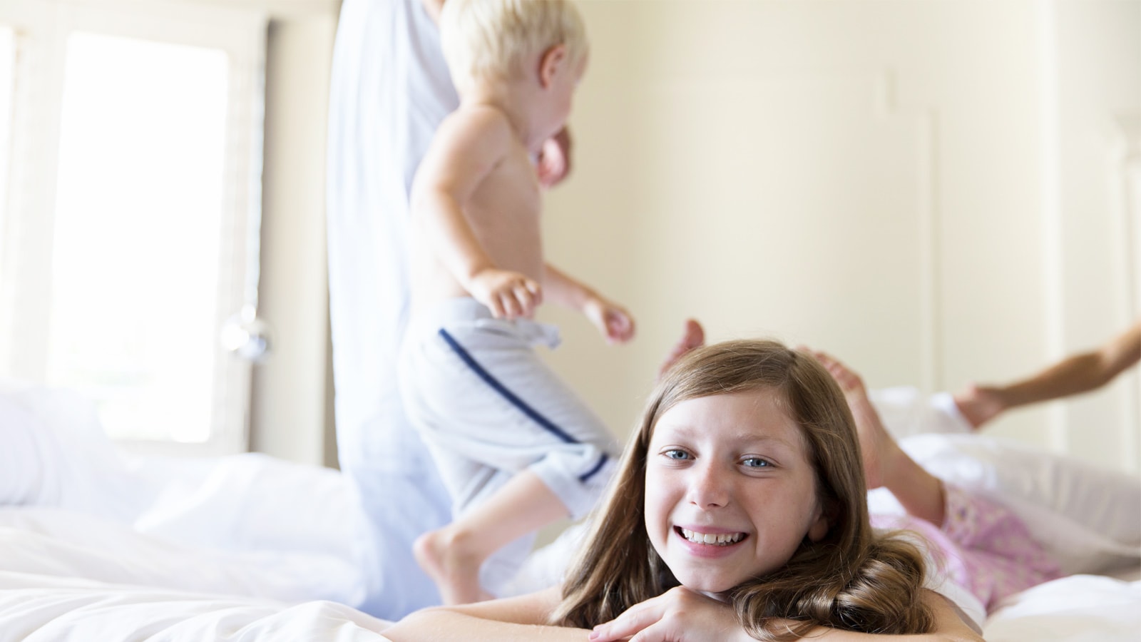 Søskende leger i seng. En lille dreng står i sengen og storesøster ligger og smiler ud mod kameraet