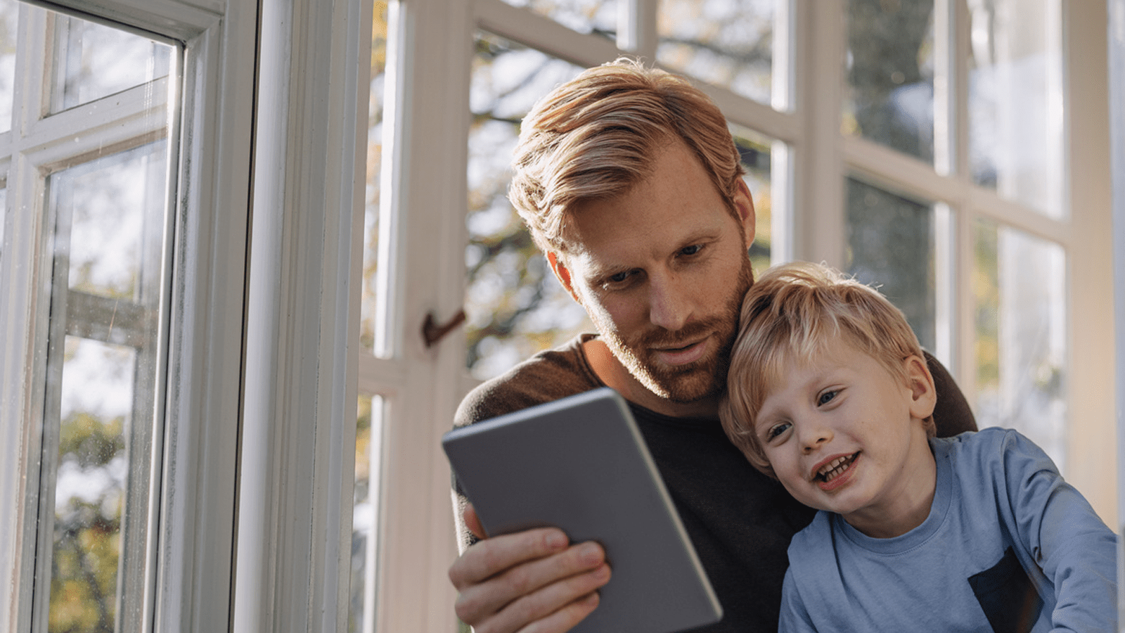 Mand og lille dreng sidder ved stort vindue med sprosser og kigger på tablet